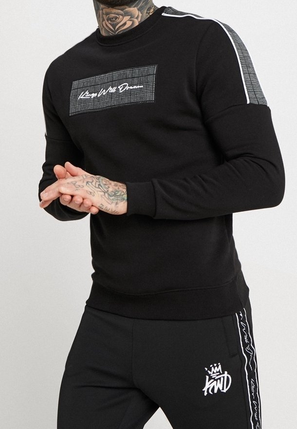 Homme portant un sweat-shirt noir et un pantalon avec un texte blanc et des patches à motifs, mains jointes, montrant des tatouages au cou et aux mains.