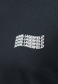 Μαύρο ύφασμα με λευκό κείμενο που επαναλαμβάνει τη φράση "KARL LAGERFELD" τέσσερις φορές σε κυματιστό μοτίβο.