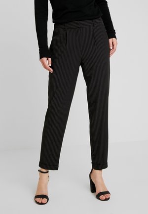 Femme portant un pantalon à pinces noir rayé, associé à des sandales à talons noirs et un haut à manches longues noir.