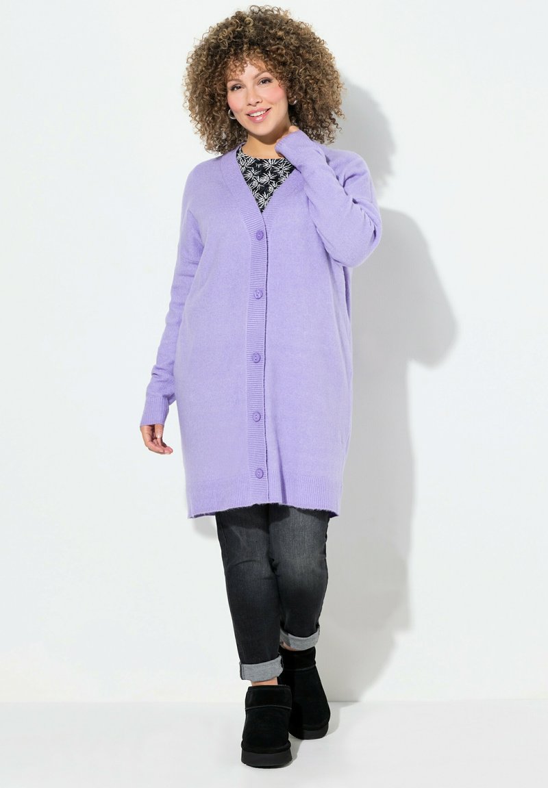 Cardigan long en lavande en tricot doux avec un devant à boutons, des poignets côtelés et une coupe décontractée, associé à un jean foncé et des bottes noires.
