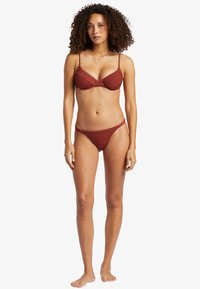 Rotes, texturiertes Bikini-Set mit Bralette-Top und dünnen Trägern sowie niedrighängenden Hosen, das ein glattes, enganliegendes Design betont.