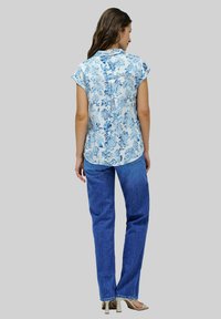 Camicia floreale blu chiaro e bianca con maniche corte, abbinata a jeans blu a gamba dritta e tacchi beige, vista da dietro.