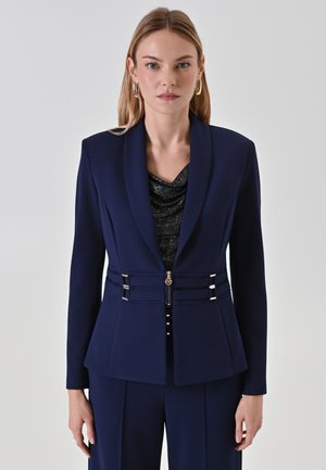 Navy blue blazer in structured fabric with a V-neckline, featuring metallic hardware accents and a tailored fit with side panel details.