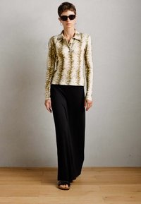 Personne portant une chemise à manches longues à motif, une jupe maxi noire, des lunettes de soleil noires et des sandales noires, se tenant sur un sol en bois contre un mur uni.