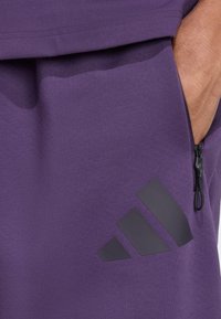 Violet sporta šorti ar fakturētu audumu, sānu kabatu ar rāvējslēdzēju un melnu diagonālu svītru logo apakšējā kreisajā pusē.