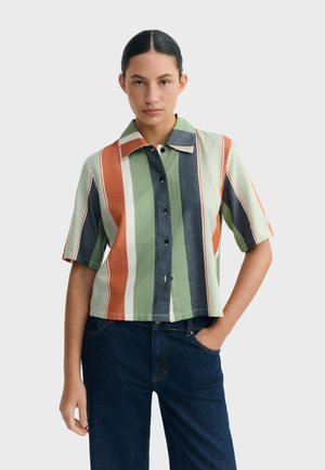 Młoda kobieta o ciemnych włosach ma na sobie koszulę na krótki rękaw z pionowymi zielonymi, pomarańczowymi, białymi i niebieskimi paskami oraz ciemnoniebieskie dżinsy.