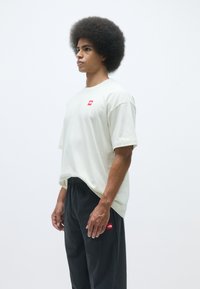 Lysegrå oversized t-shirt med korte ærmer og et lille rødt logo på brystet, parret med sorte bukser med elastisk talje, som har et rødt logo.