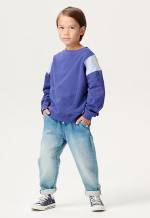 Lila Sweatshirt mit hellblauen Schulterpaneelen, kombiniert mit hellblauen, leicht baggy geschnittenen Denim-Jeans und marineblauen Sneakers. Einfaches Design, lässiger Look.