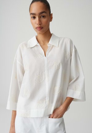Vrouw draagt een witte blouse met kraag en driekwart mouwen met schelpborduursel, hand in de zak, tegen een effen lichte achtergrond.
