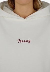 Biely mikina s textúrovaným povrchom, s bordovým vyšitým nápisom "PEGADOR" na prednej strane. Kapucňa je výrazne zobrazená.