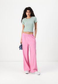 Crop top corto a maniche corte di un verde chiaro a coste, abbinato a pantaloni larghi rosa con vita elastica. La modella tiene in mano una borsa blu.