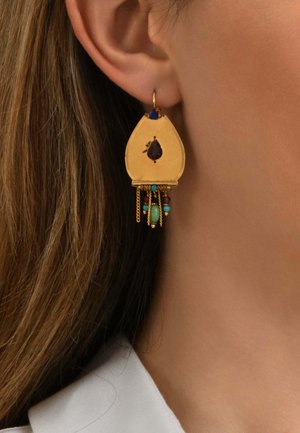 Boucle d'oreille en forme de goutte en or avec une pierre centrale sombre, des perles bleues et turquoise, et de petites chaînes suspendues portée à l'oreille d'une femme.