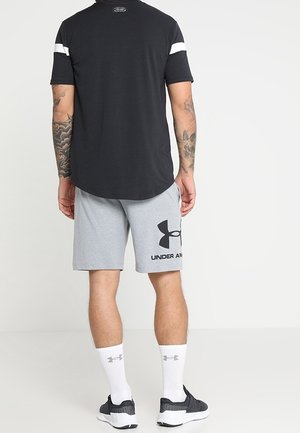 Maglietta nera a maniche corte con strisce bianche sulle spalle, abbinata a pantaloncini grigi con un grande logo nero. Calzini bianchi corti e scarpe scure completano il look.