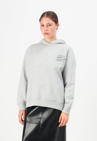 Donna che indossa una felpa grigia chiara con un piccolo design ricamato e una gonna di pelle nera, in piedi contro uno sfondo bianco.