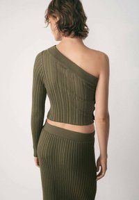 Top scurt tricotat rib și verde măsliniu, cu un mânecă lungă și un decolteu asimetric. Materialul are o textură moale și un design ajustat.