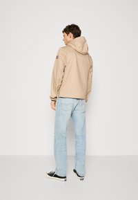 Beige hooded jacket with a smooth finish, paired with light blue jeans and black sneakers. Features a logo patch on the left sleeve.