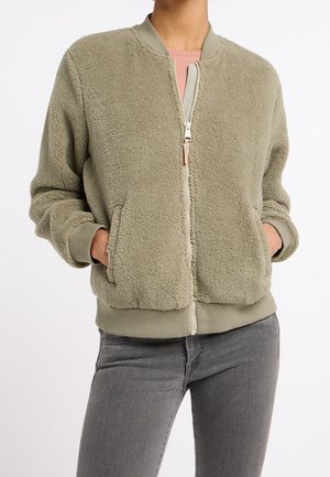 Persona que lleva una chaqueta sherpa de forro polar con cremallera de color verde oliva claro, con las manos en los bolsillos y vaqueros grises sobre un fondo blanco.