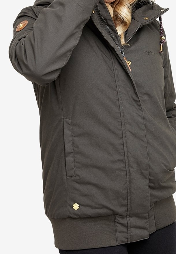 Veste matelassée vert foncé avec fermeture éclair, boutons, capuche et poches portée par une personne aux cheveux blonds.