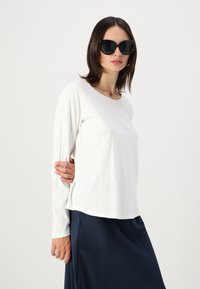 White long-sleeve top with a relaxed fit, paired with a navy skirt. Model wears large black sunglasses and has straight, dark hair.