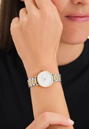 Montre-bracelet bicolore en argent et en or avec un cadran blanc, des index horaires fins et un boîtier rond, portée sur une main reposant sur un bras.