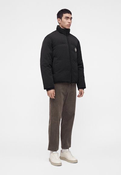 Homme debout portant une doudoune noire, un pantalon marron, des chaussettes blanches et des baskets blanches sur un fond blanc uni.