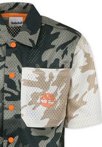 Camicia a maniche corte con motivo mimetico in tonalità di verde, beige e bianco. Presenta tessuto a rete, bottoni arancioni a contrasto e ricamo del logo.