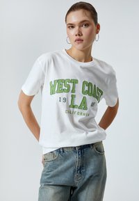 Hvid bomulds T-shirt med grøn trykt tekst "WEST COAST LA 1985 CALIFORNIA." Casual pasform, korte ærmer og blød tekstur. Båret med slidte blå denimbukser.