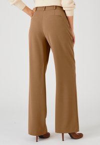 Wijdvallende broek van een warme bruine stof, met een gladde textuur, een platte voorkant en twee achterzakken. Gestyled met hoge hakken.