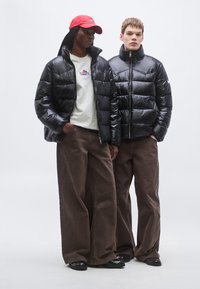 Dvaja jednotlivci nosia čierne prešívané bundy s lesklým povrchom, doplnené sivými mikinami a hnedými nohavicami s širokými nohavicami. Červená čiapka dokončuje vzhľad.