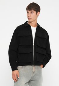Zwarte corduroy jas met een rits aan de voorkant, twee grote borstzakken en een kraag. Heeft een losse pasvorm en ribgebreide manchetten.