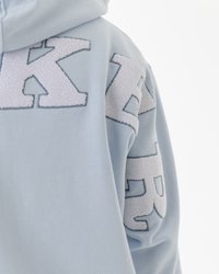 Šviesiai mėlyna gobtuvė su dideliais, išsiuvinėtais rašmenimis pūkuotos baltos tekstūros, su švelnia medžiaga ir atpalaiduojančiu įfitavimu.