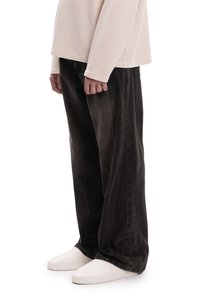 Fekete farmer széles szárú nadrág, látható varrási részletekkel, világos bézs hosszú ujjú felsővel és fehér slip-on cipővel viselve.