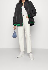 Svart satin bomberjacka med gröna detaljer och knappar, kombinerad med vita raka byxor och vita sneakers. Liten blå väska i handen.