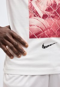 Χέρι ακουμπισμένο σε λευκό μπλουζάκι Nike με κόκκινο γραφικό μπάσκετ και μαύρο λογότυπο Nike κοντά στο τελείωμα, σε συνδυασμό με λευκό σορτς από δίχτυ.