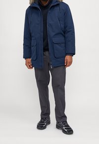 Marinblå parka med pälsfodrad huva, dragkedja och knapplås, samt två framfickor. Bärs tillsammans med grå byxor och svarta sneakers.