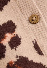Tissu tricoté beige avec un motif abstrait tacheté marron et pêche. Comprend un bouton rond foncé avec "NEXT" embossé dessus.