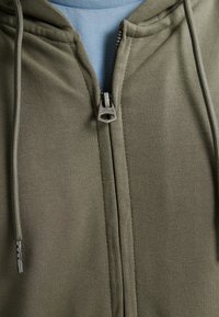 Felpa con zip verde oliva realizzata in tessuto morbido, con una cerniera in metallo e un logo, e uno strato blu chiaro sotto.