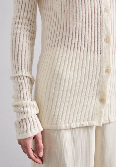Cardigan ribbed de culoare crem deschis, cu dungi subțiri maro, având închidere cu nasturi și o textură moale. Mânicile cu manșetă se extind până la încheieturi.