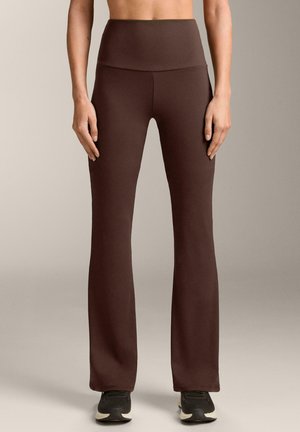 Vrouw die donkerbruine, hoge taille wijde leggings en zwarte sneakers draagt, staand tegen een neutrale achtergrond.