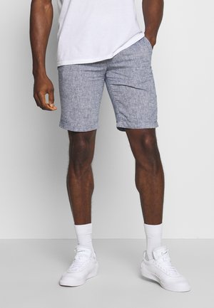 Męskie szorty w niebieskim melanżu o luźnym kroju, do kolan, wykonane z lekkiego materiału. W komplecie z białymi sneakersami i skarpetami do kostek.