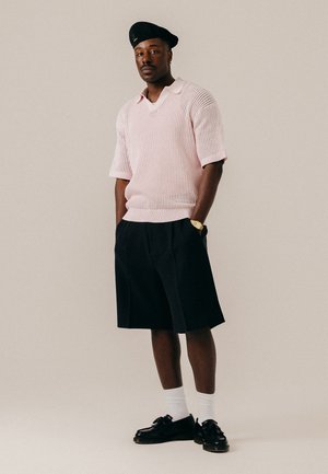 Homme portant un béret noir, un pull en maille rose clair à manches courtes, un short noir au genou, des chaussettes blanches et des mocassins noirs, debout les mains dans les poches.