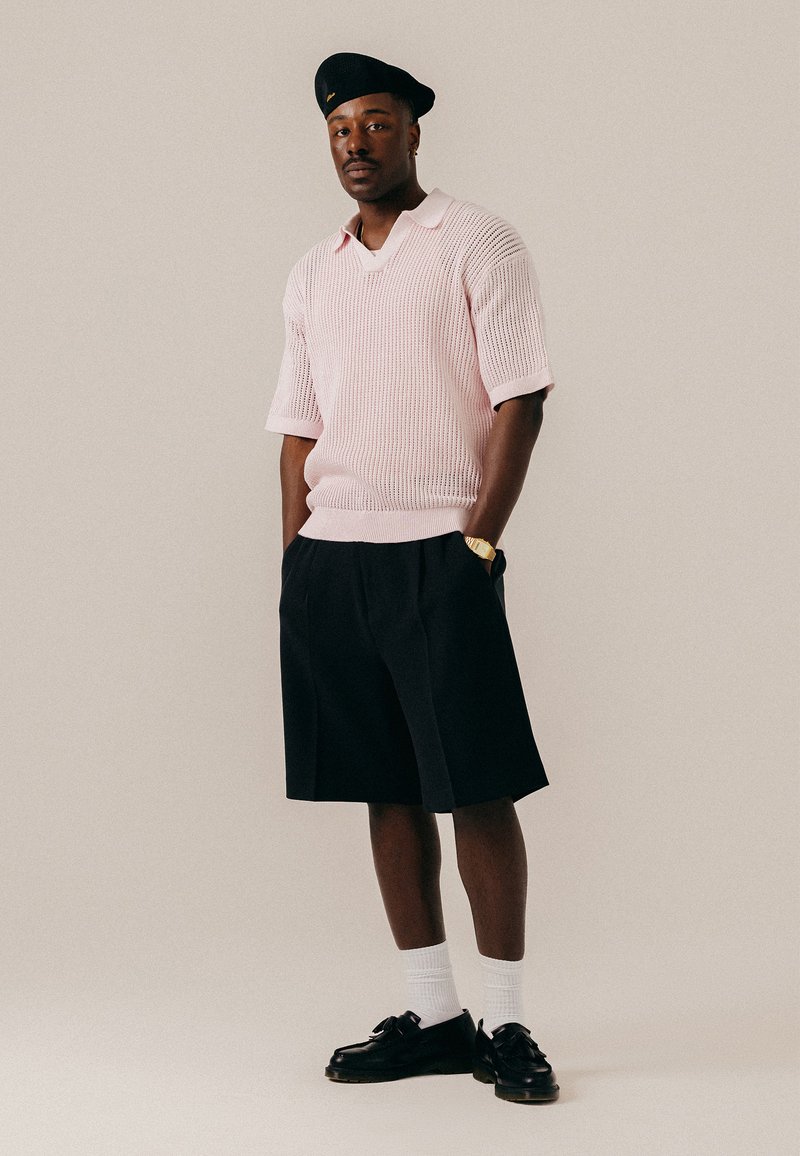 Homme portant un béret noir, un pull en maille rose clair à manches courtes, un short noir au genou, des chaussettes blanches et des mocassins noirs, debout les mains dans les poches.