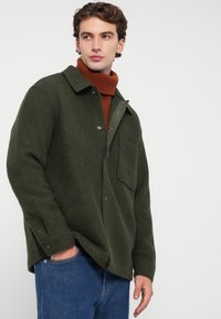 Vihreä villasekoiteovershirt, jossa on kaulus, etutasku ja nepparit, kerroksittain ruskean poolopaidan päällä ja yhdistettynä sinisiin farkkuihin.