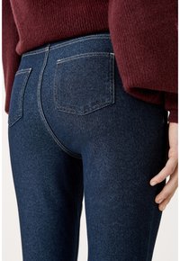 Tmavě modré džíny z denimu s přiléhavým střihem, které mají dvě zadní kapsy a kontrastní šití. Textura vypadá hladce a mírně elasticky.