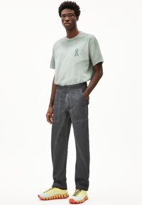 Grijze katoenbroek met contrasterende stiksels, gecombineerd met een lichtgroen T-shirt met een logo. Felgele en oranje sneakers maken de look compleet.
