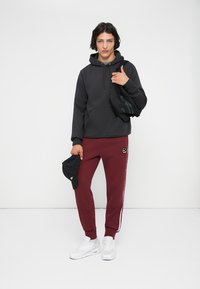 Sweatshirt preta com capuz e um bolso frontal, combinado com calças de jogging bordô com riscas laterais e um logótipo, ténis brancos e um boné preto.