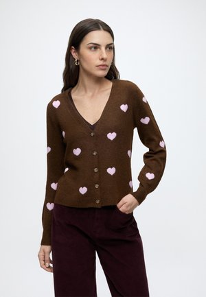 Femme aux longs cheveux foncés portant un cardigan marron boutonné avec des cœurs roses et un pantalon sombre, debout avec une main dans la poche.