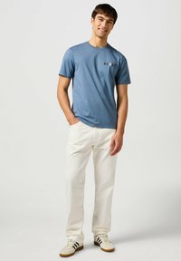  camiseta de algodón azul con mangas cortas que presenta el texto "HOWDY" en rojo y blanco. Combinada con unos pantalones vaqueros blancos y zapatillas beige.