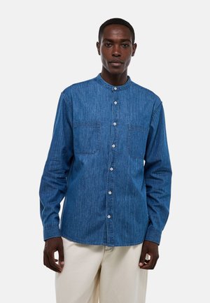 Chemise en denim bleu moyen avec un col mandarin, des manches longues, deux poches poitrine et un boutonnage à l'avant. Le tissu a une finition texturée.