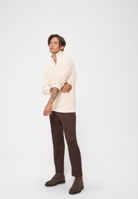 Miękki sweter na zamek błyskawiczny o prążkowanej fakturze, w połączeniu z wąskimi brązowymi spodniami i brązowymi zamszowymi botkami. Modelka pozuje z założonymi rękami.